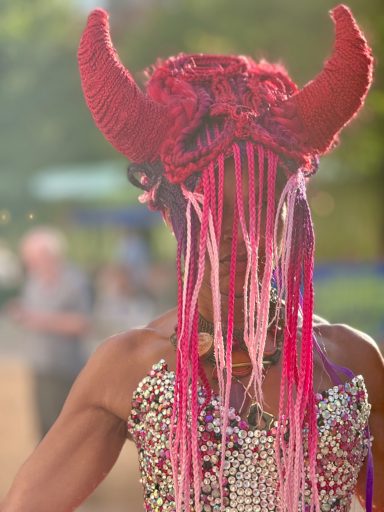 Person with red horns and colorful hair, wearing a beaded outfit, facing away.