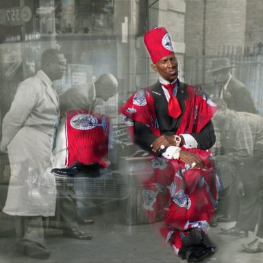 A man in a red patterned outfit and hat sits confidently in a blurred vintage urban setting in the background.