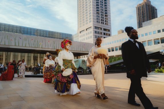 People in colorful traditional attire walking in a city plaza during an outdoor event.