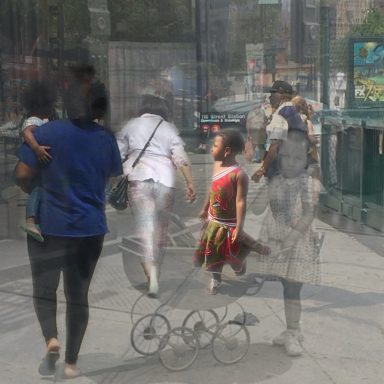 People walk in an urban area, with reflections creating a layered effect, with a little girl in a red dressed centered.