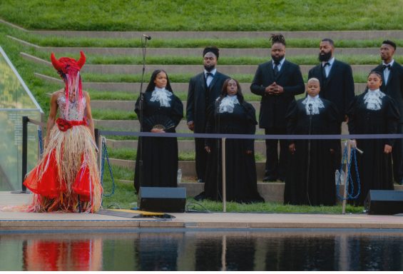 A performer in a red costume stands in front of a choir, set by a poolside stage.