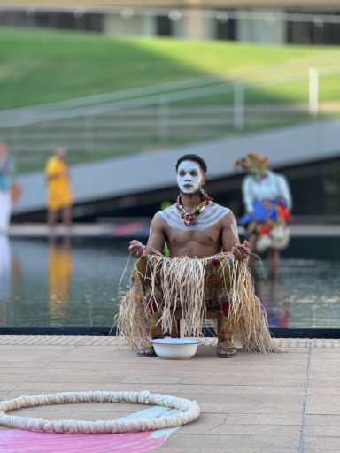 A performer in traditional attire sits by a water feature, adorned with decorations.