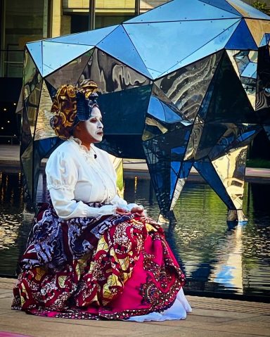 A performer in traditional attire sits beside a modern geometric sculpture.