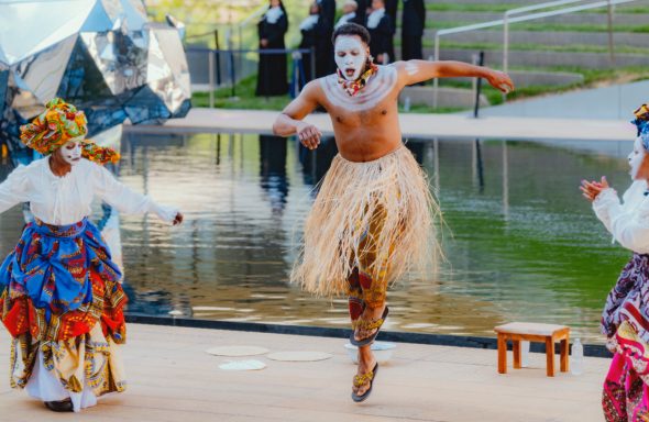 A performer in traditional attire dances by a water feature, accompanied by two others.