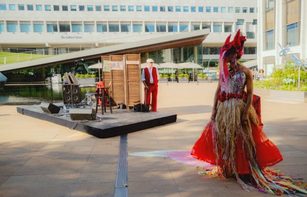 A performer in vibrant attire stands on an outdoor stage with musicians in the background.