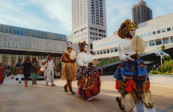 People dressed in traditional costumes parade through an urban plaza.