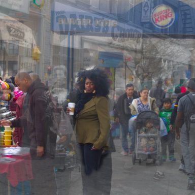 A woman in a green jacket stands in a bustling street scene with people, shops, and colorful displays.