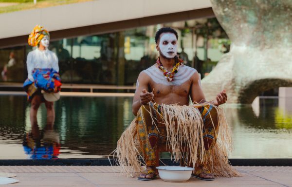 Person in traditional attire with face paint and grass skirt, performing in front of a water feature.