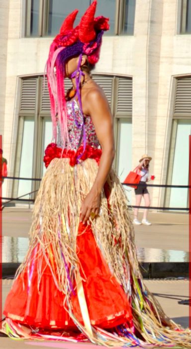 Person in vibrant costume with colorful accessories and woven skirt, striking a bold pose.