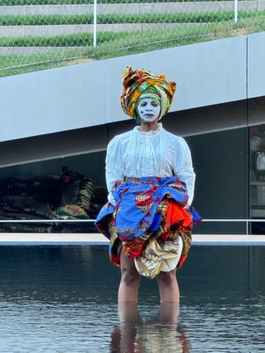 Person standing in water, wearing a colorful outfit and head wrap, is painted white.