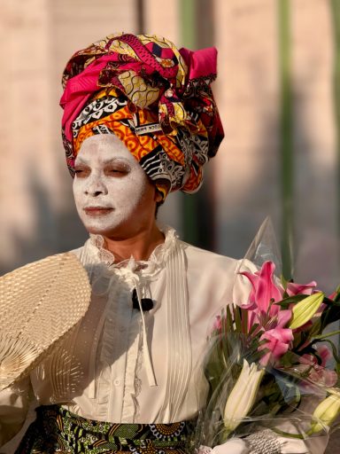 A woman in traditional attire with a colorful head wrap holds a bouquet of flowers.