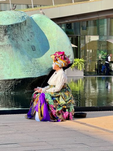 A person in a colorful traditional outfit sits near a modern fountain.