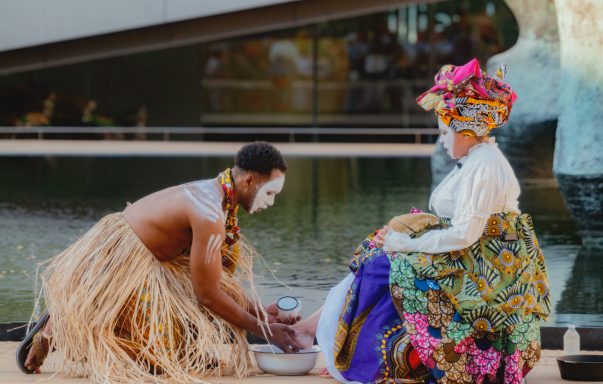 A man in traditional attire performs a ritual beside a woman in colorful clothing.