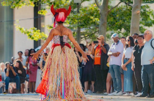 A performer in a vibrant costume with horns and a straw skirt dances in front of an audience.