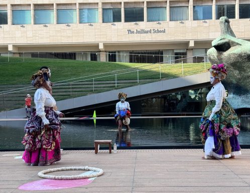 Two women in colorful traditional dresses perform near a water feature with a sculpture.