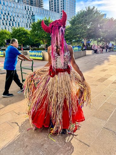 Person in a colorful, fringed dress with horns, standing in an outdoor setting.