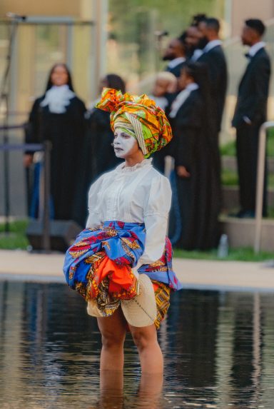 Person in colorful attire and face paint standing in water, with others in formal wear nearby.