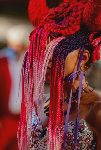 Person with vibrant red and pink braided hair and elaborate horns, dressed in sparkly attire.