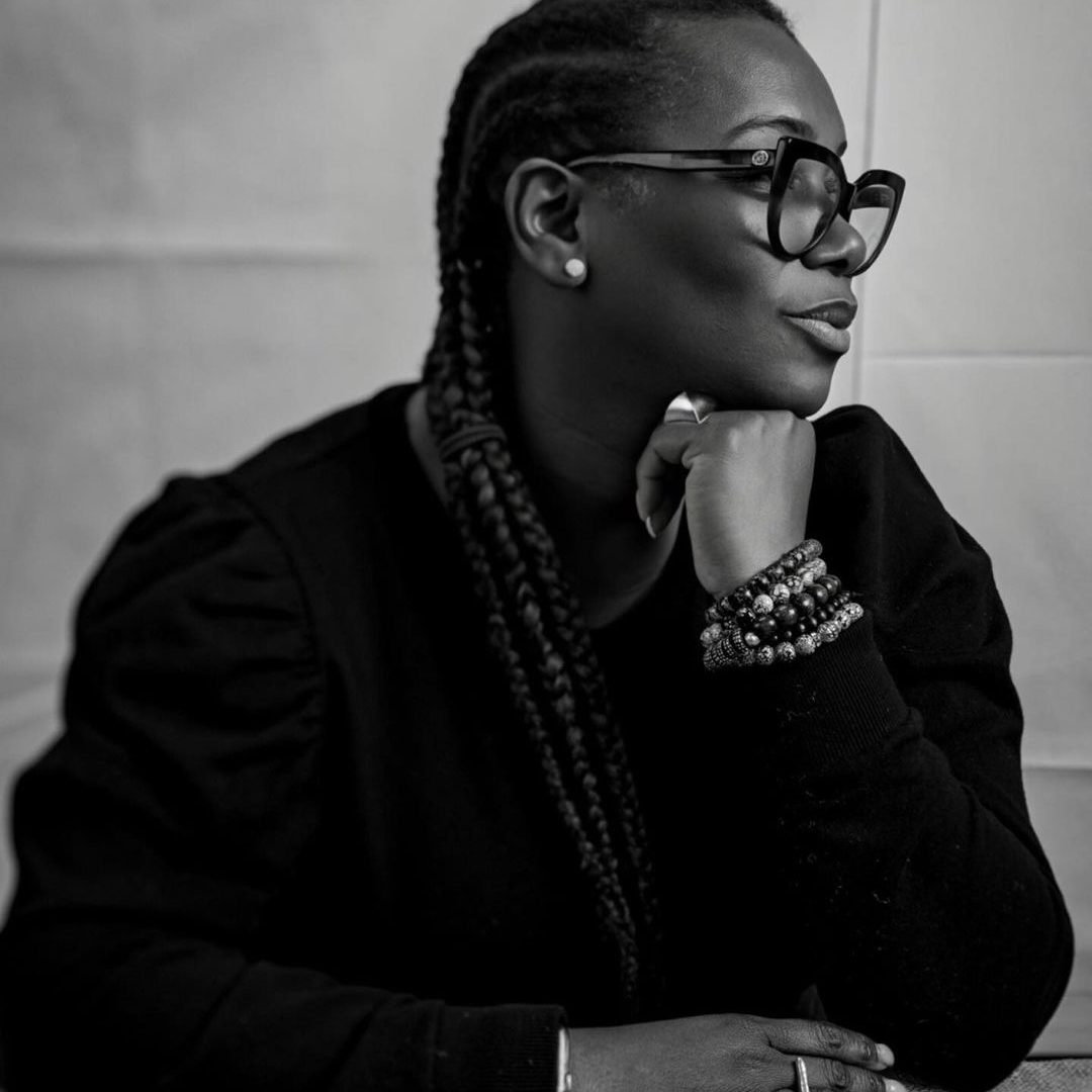 A woman with braided hair and glasses poses thoughtfully with her hand on her chin.