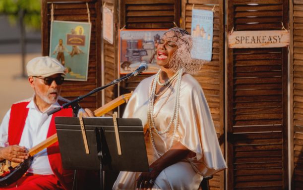 Musicians performing outdoors; a man plays a string instrument while a woman sings.