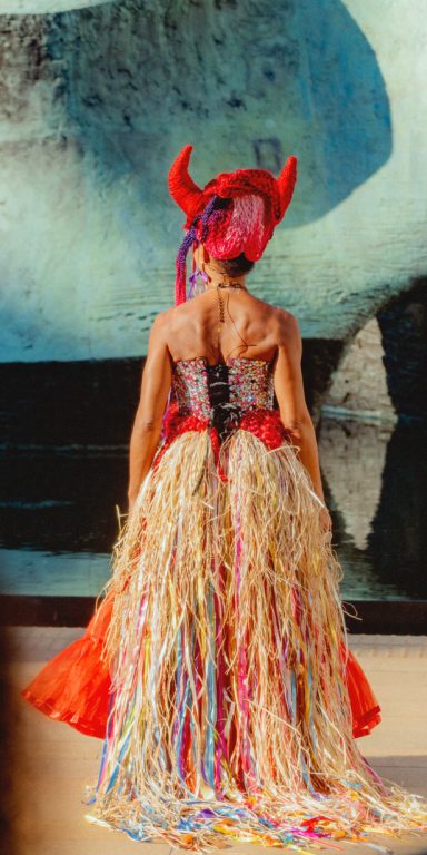 Woman in a colorful dress with a headdress, standing against an abstract background.