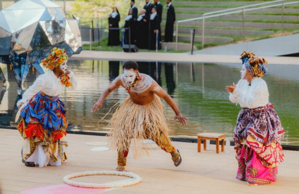 Three performers in traditional costumes engage in a lively dance near water.