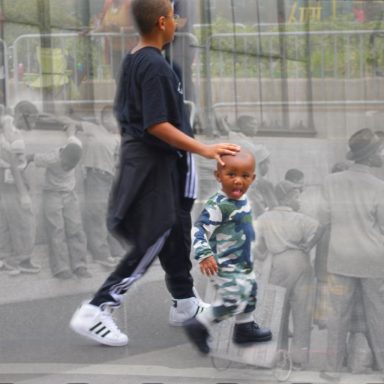 Two children walking together, the taller one has a hand, on the smaller child's head while with blurred figures in the background.