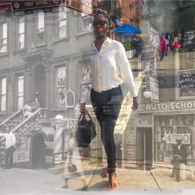 A woman in a white blouse and sunglasses walks past a historic urban scene.